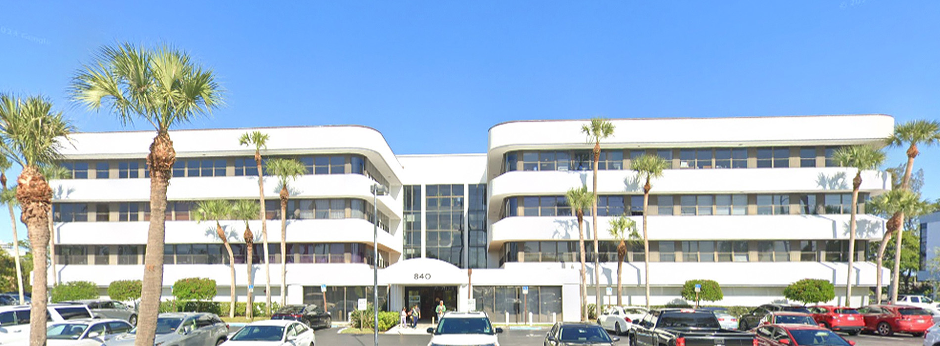 The image shows a multi-story building with a modern architectural design, featuring white walls, large windows, and palm trees in the foreground.