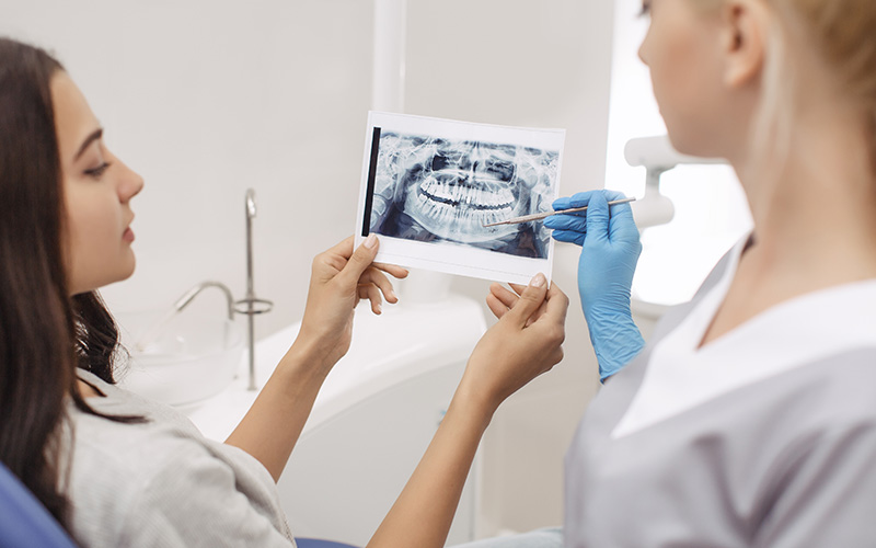 In the image, a dental hygienist is holding up an X-ray of a patient s teeth for examination, while a woman in a blue shirt watches the process.