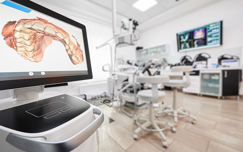 The image shows a modern dental office with a large digital display screen in the foreground, featuring an animated 3D model of a human mouth and teeth. In the background, there is a dental chair, various pieces of dental equipment, and a professional setting with a clean aesthetic.
